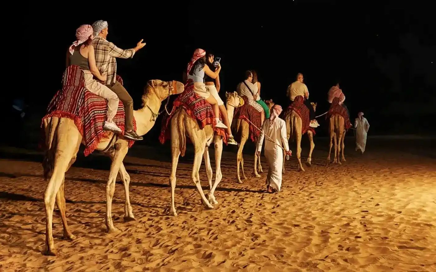 Visitors riding a line of camels beside a desert camp at night