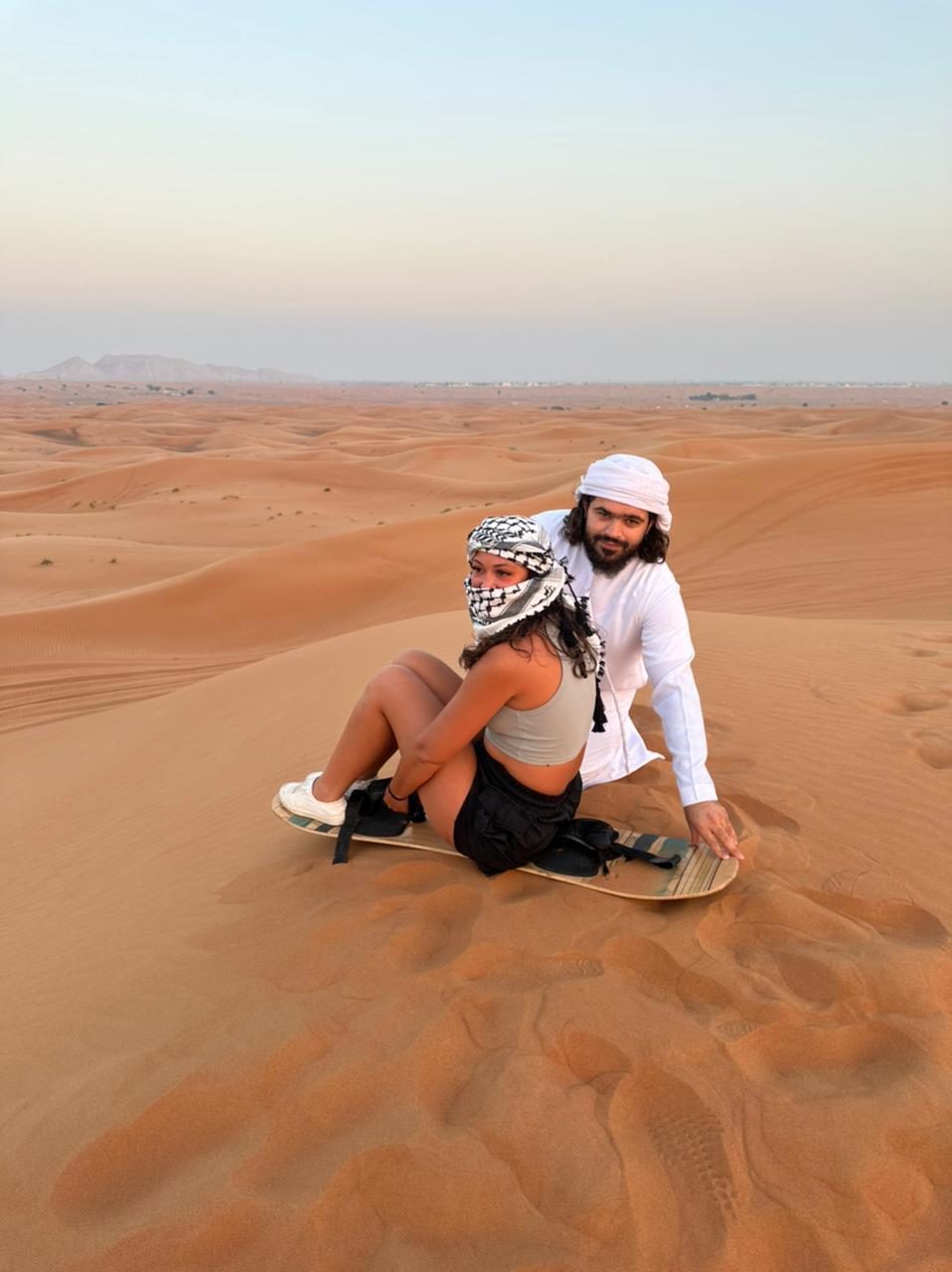 Visitors seated on a sandboard at the top of a desert dune
